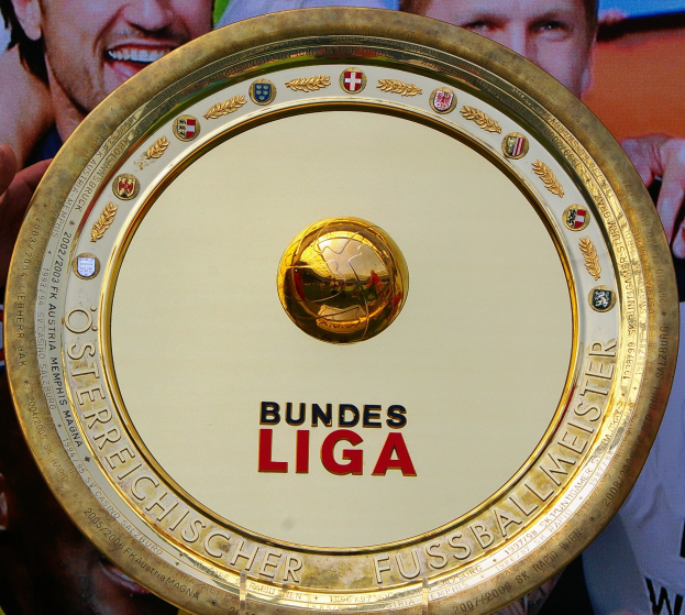 Ein Mann hält ein goldenes Schild mit der Aufschrift 'Bundesliga', im Hintergrund eine Fahne mit drei Personen.