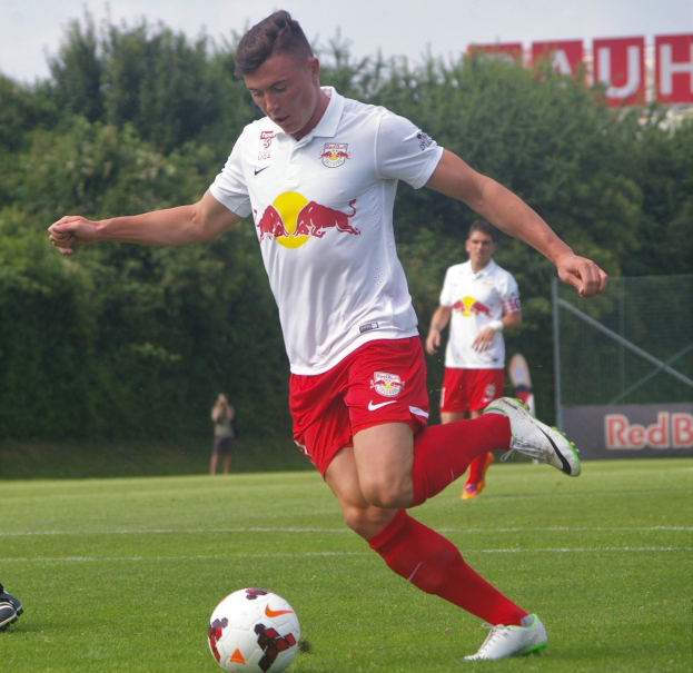 Ein professioneller Fussballspieler tritt einen Ball auf einem Rasenfeld mit Bäumen und einem klaren blauen Himmel im Hintergrund, in der Nähe eines Schildes mit der Aufschrift "RB Leipzig."