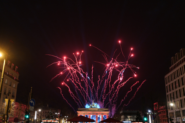 Eine belebte Stadtstraße bei Nacht mit vielen Menschen, Fahrzeugen, Zelten und Gebäuden, mit einem farbenfrohen Feuerwerk am Himmel im Hintergrund.