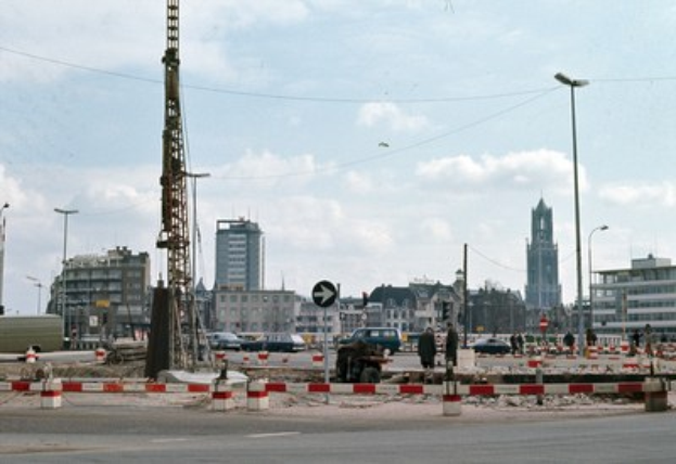 Bauplatz mit einem zentralen Turm, umgeben von Gebäuden, Straßeninfrastruktur, Fahrzeugen und einem bewölkten Himmel.