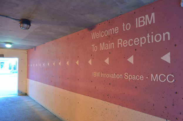 Eine Wand mit dem Text "Willkommen bei IBM", beleuchtet von Deckenlampen, mit einer Tür im Hintergrund, die "IBM Innovation Space MCC" bezeichnet.