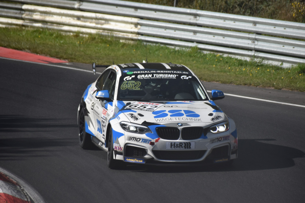 BMW M4 DTM Rennauto auf einer Rennstrecke mit Gras, Zaun und Pflanzen, mit Text und Zahlen auf der Karosserie.