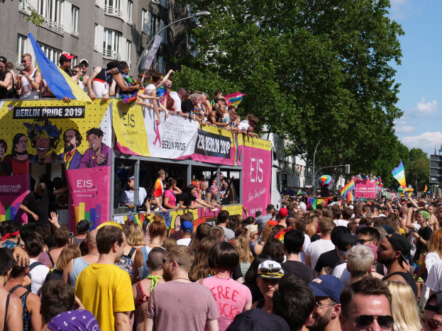 Eine große Menschenmenge marschiert auf der Straße der Berlin Pride 2019, viele tragen Mützen und Schutzbrillen, halten Fahnen, mit Bannern und Gebäuden im Hintergrund bei bewölktem Himmel.