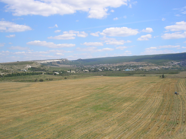 Ein Traktor auf einem kultivierten Feld umgeben von Bäumen, Gebäuden, Hügeln und einer bewölkten Himmel