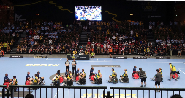 Eine Gruppe von Rollstuhlfahrern bei den Invictus Games Düsseldorf 2020 auf einem von einem Geländer umgebenen Platz, mit einer großen Zuschauermenge im Hintergrund und einer Treppe, die zu einem Bildschirm führt.