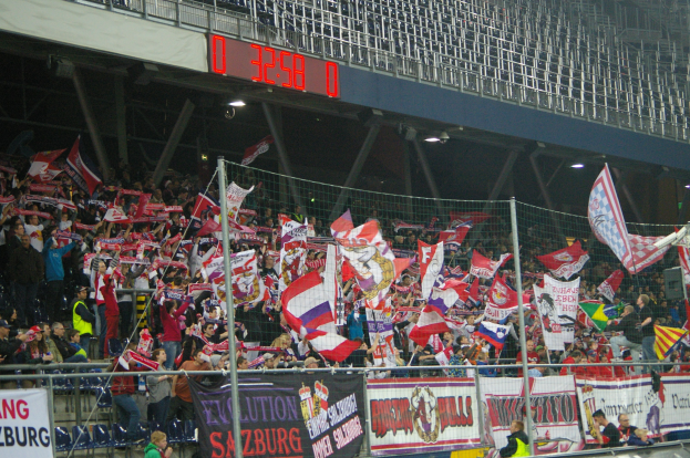 Eine große Menschenmenge in einem hell erleuchteten Stadion schwenkt Fahnen und Banner mit Text, getrennt durch ein Netz von einer digitalen Anzeigetafel oben.