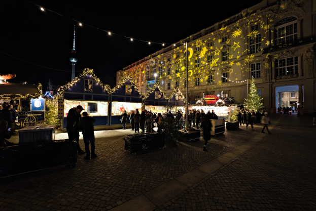 Ein geschäftiger Weihnachtsmarkt in Berlin, Deutschland mit Menschen um dekorierte Stände, festliche Lichter und Gebäude im Hintergrund unter einem dunklen Himmel.