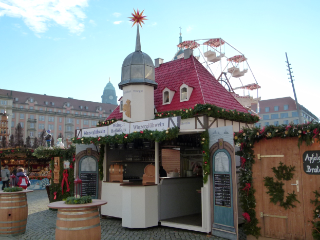 Kleines Gebäude mit rotem Dach und Riesenrad davor, umgeben von Festobjekten und Menschen, mit Gebäuden, Bäumen und bewölktem Himmel im Hintergrund.