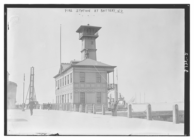Schwarzes und weißes Foto einer Feuerwache an der Battery, New York, mit Menschen, Pfählen, einem Zaun, Booten auf dem Wasser und einem bewölkten Himmel.