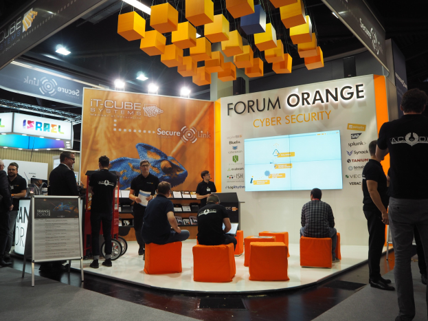 Eine Gruppe von Menschen an der IT-Cube Systems Forum Orange Cyber Security Theke, einige sitzen auf Stühlen, andere stehen, umgeben von Anzeigen, Regalen, Schildern und Deckenleuchten.