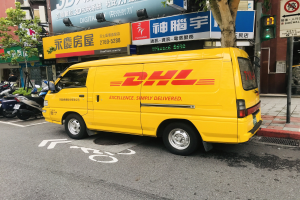 Gelber DHL-Lieferwagen geparkt am Straßenrand, umgeben von Gebäuden, Bäumen, Schildern, Strommasten, Ampeln und einem Fußgänger auf dem Gehweg.