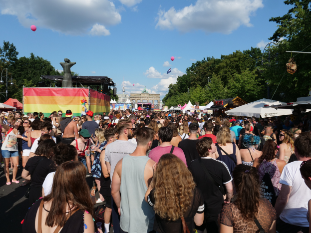 Eine große Menschenmenge, die eine Straße mit Zelten, Bäumen, Pfählen, Lichtern und einer Statue gesäumt, mit Gebäuden und einem bewölkten Himmel mit Ballons im Hintergrund auf dem Berlin Pride Festival.
