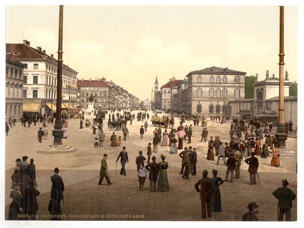 Eine belebte Münchner StraßenSzene aus den frühen 1900er Jahren mit Menschen, die spazieren gehen, einige mit Schirmen, und einer Kutsche im Vordergrund, Gebäuden, Bäumen und Polen im Hintergrund und Text am unteren Rand des Bildes.