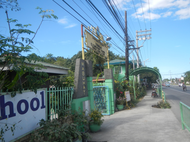 Einfahrt zu einer Schule mit einem "Schule"-Schild am Straßenrand, mit einem Torbogen, einem Tor, einer Namensplatte, Fahrzeugen, Pflanzen, Bäumen, Strommasten mit Drähten und einem bewölkten Himmel.