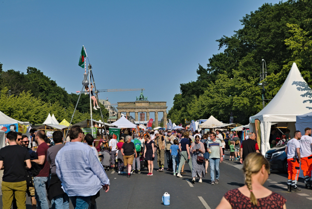 Eine Menschenmenge, die eine Straße entlanggeht, die von Zelten, Fahrzeugen und Bäumen gesäumt ist, mit einem Bogen und einem klaren blauen Himmel im Hintergrund und Fahnenmasten auf der linken Seite, die wahrscheinlich das Oktoberfest in München, Deutschland, anzeigen.