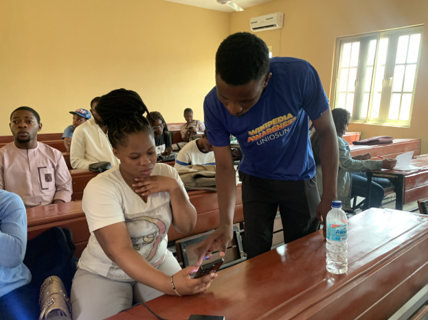 Eine Klasse mit Schülern an Tischen, ein Mann mit einem Mobiltelefon in der Hand, Flaschen und andere Gegenstände auf den Tischen und eine Klimaanlage an der Wand mit einem Fenster im Hintergrund.