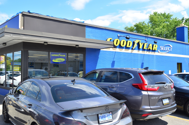 Ein Goodyear Select Autohaus mit mehreren geparkten Autos davor, das ein Gebäude mit Text darauf und Bäume mit einem bewölkten Himmel im Hintergrund zeigt.