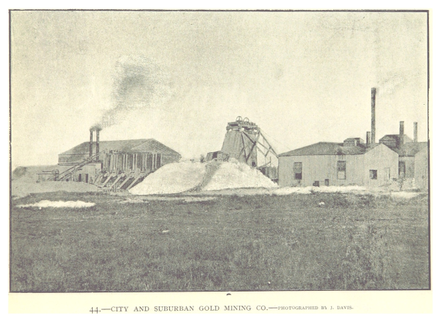 Schwarz-weißes Foto einer Stadt und eines suburbanen Goldbergbauunternehmens mit Gebäuden, Polen und Rauch im Hintergrund, mit Text unten.