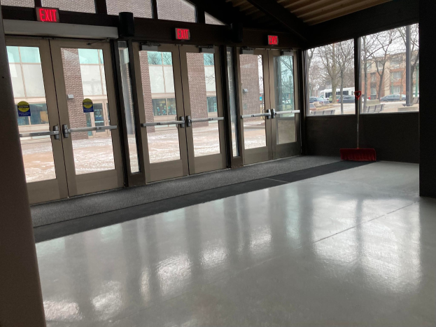 Großer Raum mit hoher Decke, vielen Fenstern und Türen, mit einem Besen auf dem Boden, Schildtafeln mit Text, einem sichtbaren Dach, umliegenden Bäumen, Fahrzeugen auf dem Boden, benachbarten Gebäuden mit Fenstern und einem bewölkten Himmel, was auf einen gewerblichen Bodenreinigungsdienst hinweist.