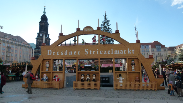 Ein lebendiger Weihnachtsmarkt in Dresden, Deutschland, mit Menschen, die herumlaufen, einem Bogen mit Text, Bäumen, Gebäuden mit Fenstern und einem bewölkten Himmel.