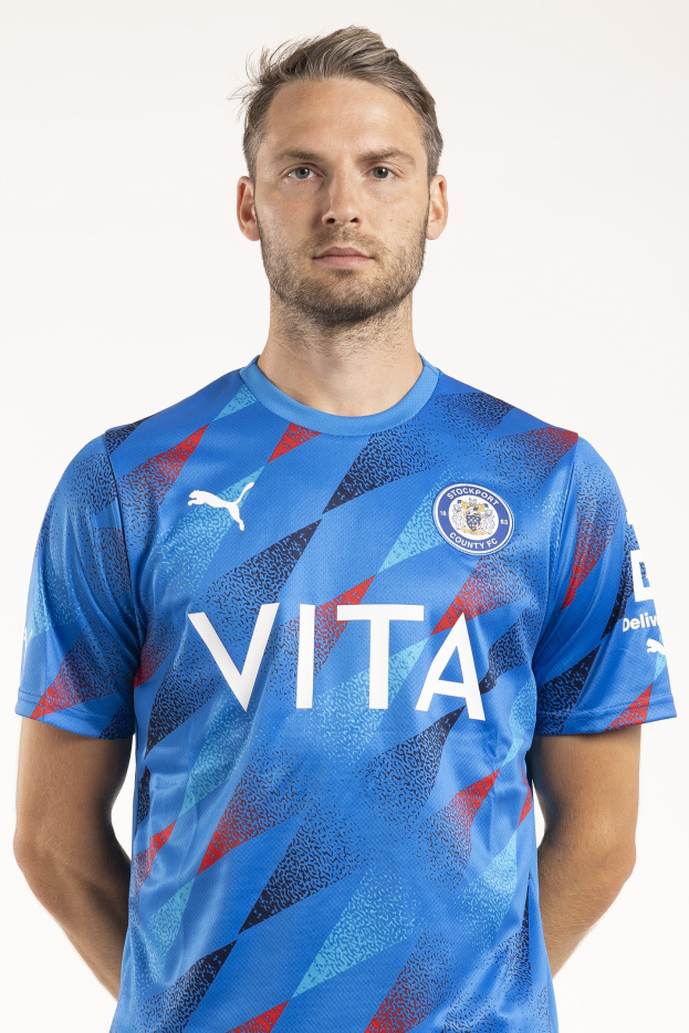 Ein Mann steht vor einem weißen Hintergrund und trägt ein blaues Leicester City Away-Shirt aus der Saison 2018-19, das das Team-Logo und Text auf der Vorderseite aufweist.