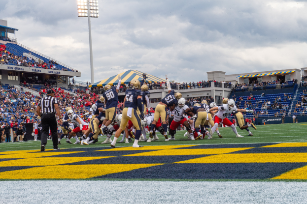 Ein Fußballspiel wird in einem Stadion gespielt, mit Spielern in Sportkleidung und Helmen im Vordergrund und Zuschauern, Geländern, Tafeln mit Text, Laternen und einem bewölkten Himmel im Hintergrund, mit Text unten auf dem Bild.