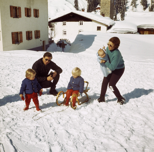 Familie von vier Personen beim Schneespiel, mit einem Mann auf einem Schlitten, einer Frau mit einem Baby auf dem Arm und zwei Kindern in der Nähe, Häuser und Bäume im Hintergrund unter einem klaren blauen Himmel.
