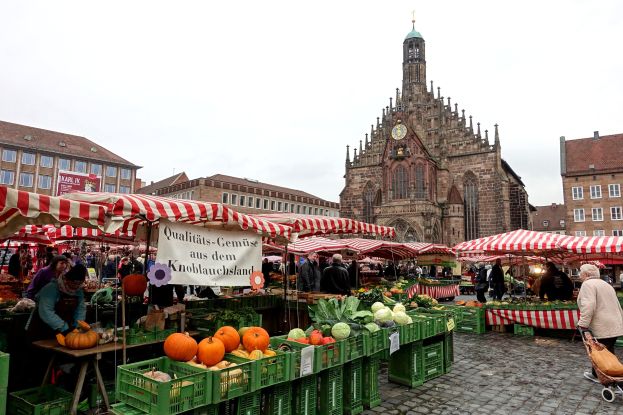 Ein belebter Markt in Nürnberg, Deutschland, mit farbenfrohen Obst- und Gemüseständen, Menschen mit Taschen, Zelten und einem Uhrenturm im Hintergrund unter einem sichtbaren Himmel.