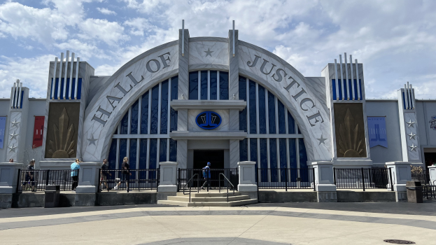 Eingang zum Hall of Justice im Disney's Hollywood Studios mit einer Gruppe von Menschen davor, Text und Banner an dem Gebäude, bewölkter Himmel.