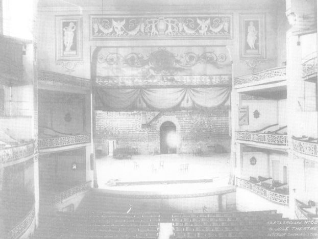 Schwarz-weiß-Foto des Theatre Royal London Auditoriums, das eine Bühne mit Stühlen, Geländern, skulptierten Wänden und einem Vorhang zeigt, mit Text am unteren Rand.