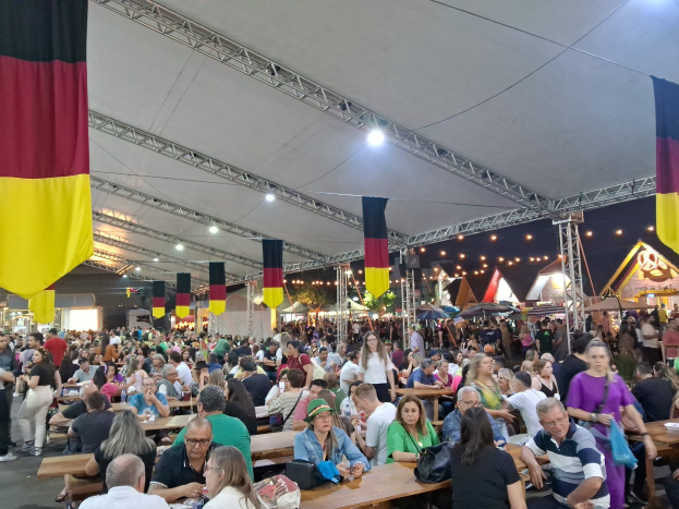 Große Gruppe von Menschen sitzt an Tischen unter einem Zelt auf dem Oktoberfest in München, Deutschland, mit Taschen und Gegenständen auf den Tischen und festlicher Dekoration im Hintergrund.