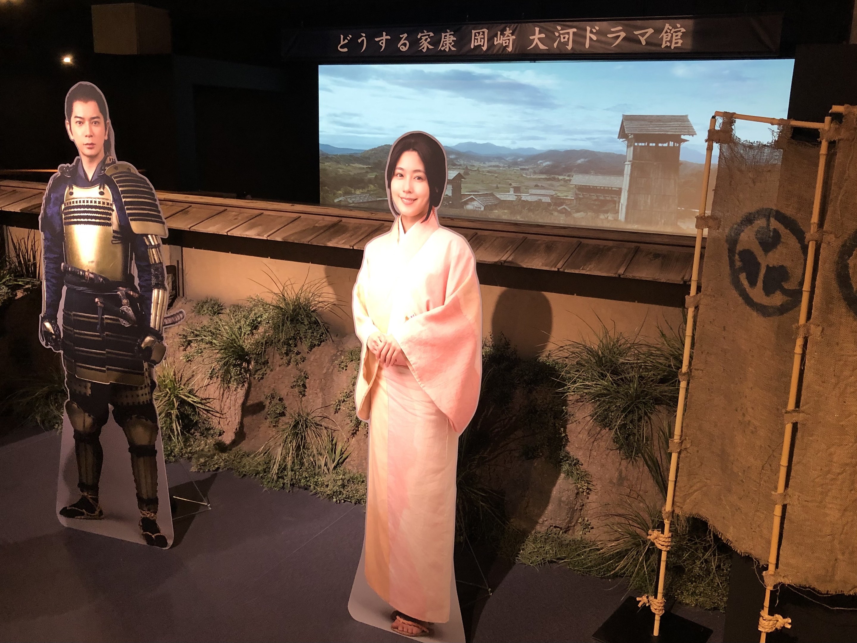 Eine Frau in einem rosa Kimono und ein Mann in einem Samurai-Kostüm, die gemeinsam auf einem japanischen Filmset mit Pflanzen, einem Bildschirm und Scheinwerfern im Hintergrund lächeln.