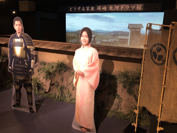 Eine Frau in einem rosa Kimono und ein Mann in einem Samurai-Kostüm, die gemeinsam auf einem japanischen Filmset mit Pflanzen, einem Bildschirm und Scheinwerfern im Hintergrund lächeln.