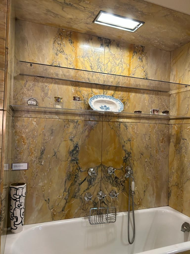 Bad mit Wanne und Dusche, mit einem Hahn, Ständer, Vasen und Gegenständen auf Regalen; Wände haben Marmorfliesen und Decke hat Beleuchtung.