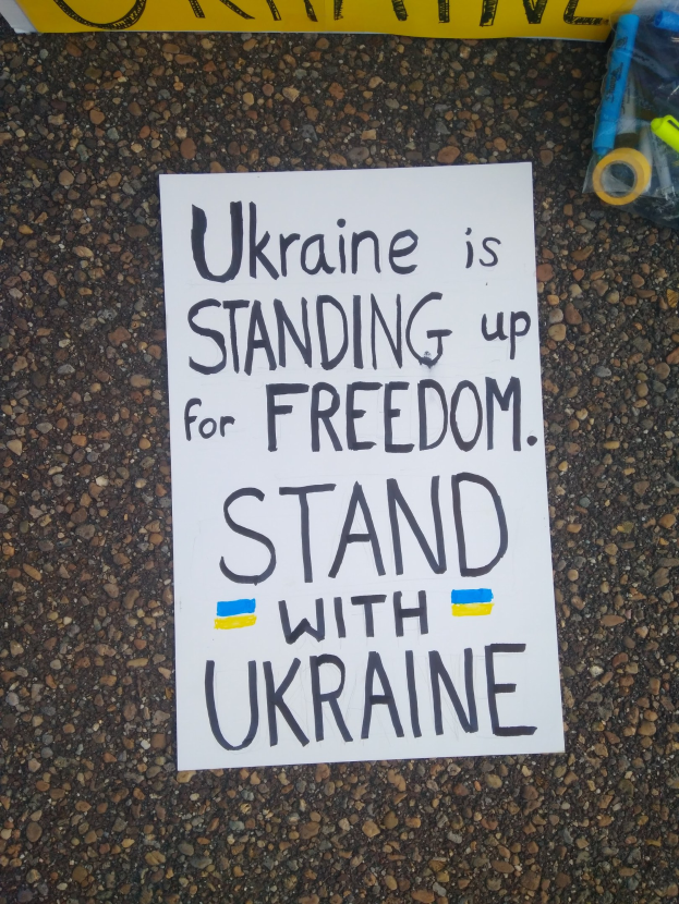 Ein weißer Zettel mit dem Text "Ukraine steht für Freiheit ein, steht mit Ukraine" liegt vor einer gelben Fahne auf dem Boden. Rechts daneben befindet sich eine Plastikabdeckung mit verschiedenen Gegenständen.