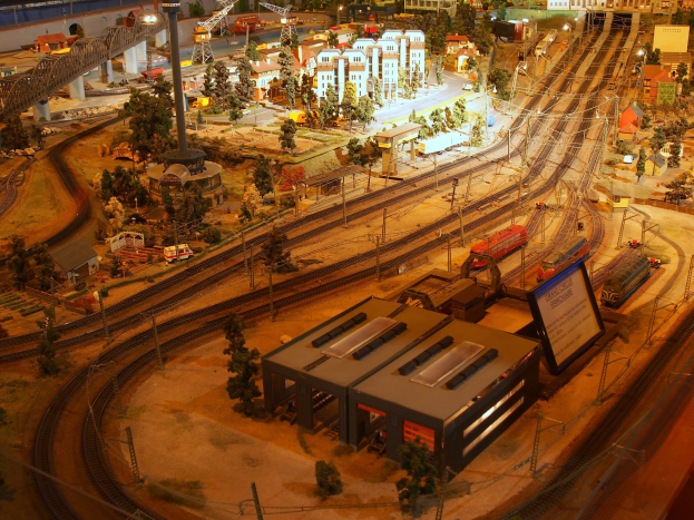 Eine detaillierte Modellbahnausstellung in einem Museum, die Gebäude, Bäume, Eisenbahnschienen, Fahrzeuge, Pfosten, Lichter und Platten zeigt, die eine belebte Stadtlandschaft nachahmen.