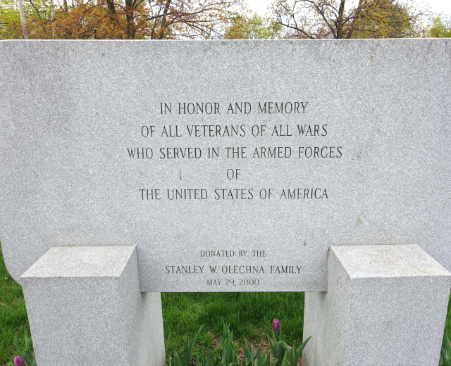 Ein Gedenkstein mit der Inschrift "In Ehren und Erinnerung an alle Veteranen aller Kriege, die in den Streitkräften der Vereinigten Staaten von Amerika dienten" umgeben von Pflanzen, Blumen, Gras, Bäumen und einem bewölkten Himmel.