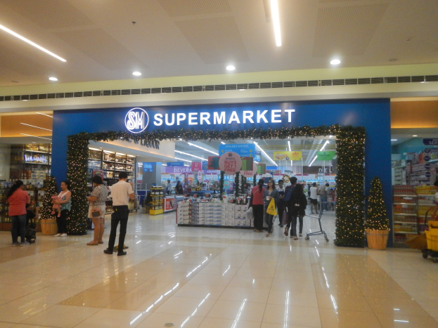 Eine Gruppe von Menschen, die vor einem Supermarkt stehen, einige halten Einkaufstaschen, mit einem Einkaufswagen, Topfpflanze, Regalen, Schildern und Deckenleuchten.