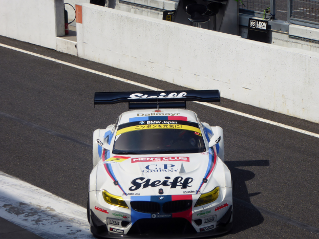 Ein BMW M6 GT3 Rennwagen auf einer Rennstrecke mit einer Wand, einem Maschendrahtzaun und anderen Objekten im Hintergrund.