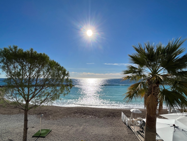 Ein sonniger Strand mit Palmen, Sonnenschirmen und üppiger Vegetation, vor einem hellblauen Himmel mit strahlender Sonne im Hintergrund.