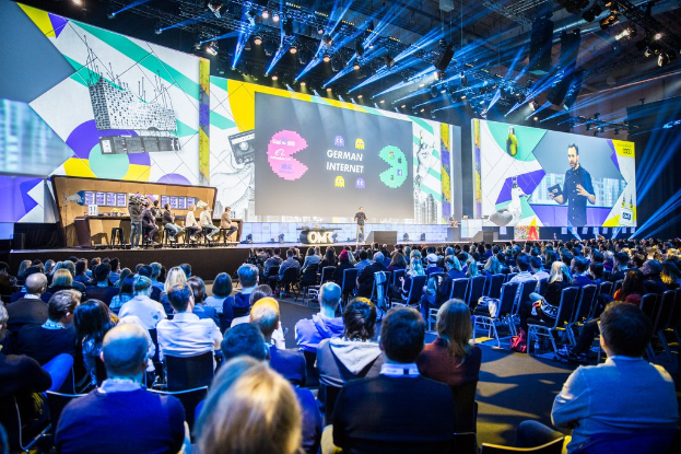 Eine große Menschenmenge, die in Stühlen vor einer Bühne sitzt, mit einigen Individuen, die auf dem Podium im Vordergrund stehen, einem Tisch mit verschiedenen Gegenständen links und Bildschirmen und Lichtern im Hintergrund auf der Deutschen Internet-Konferenz 2019.