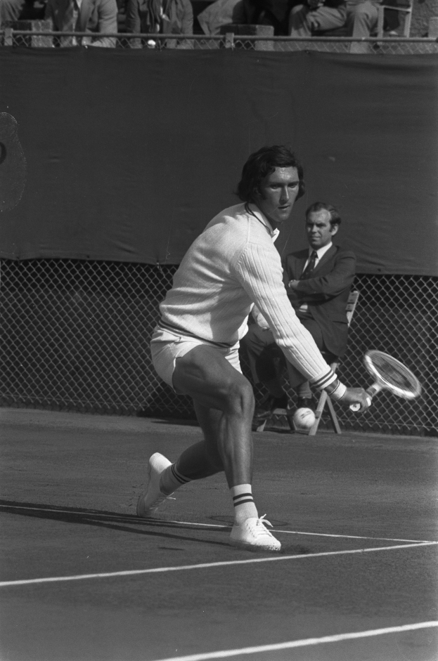 Ein professioneller Tennisspieler in Sportschuhen schlägt mit einem Schläger einen Ball, mit Maschendrahtzaun und Zuschauern im Hintergrund, die auf Stühlen sitzen.