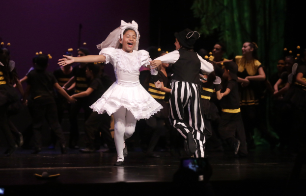 Ein junges Mädchen in einem weißen Kleid und ein Mann in einem schwarz-weißen gestreiften Anzug tanzen auf einer Bühne mit kostümierten Zuschauern, einem grünen Vorhang und einer violetten Wand.