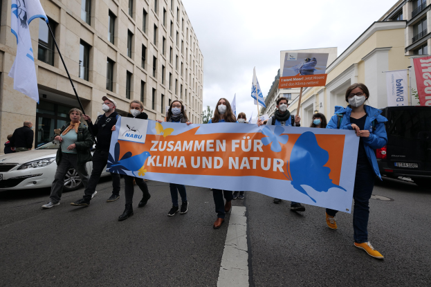 Eine Gruppe von Menschen mit Masken, die eine Schilder tragen, die "Zusammen für Klima und Natur" lautet, gehen eine Straße entlang, mit Fahrzeugen, Gebäuden, Bäumen und einem klaren blauen Himmel im Hintergrund.