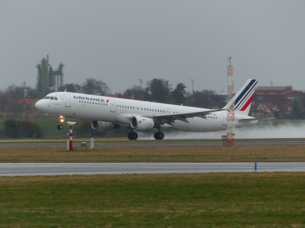 Air France Airbus A320-200 beim Start vom Pariser Charles-de-Gaulle-Flughafen, umgeben von grünem Gras und Bäumen, mit Gebäuden und Türmen im Hintergrund unter einem klaren blauen Himmel.