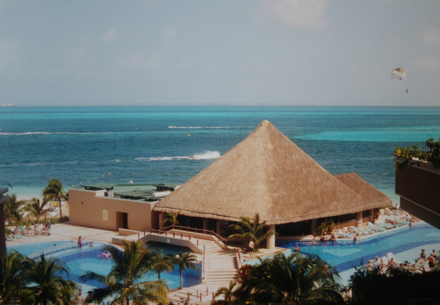 Resort mit großem Swimmingpool, umgeben von Palmen, Hütten und Menschen, mit einem Ozean, einer bewölkten Himmel und einem Fallschirm im Hintergrund.