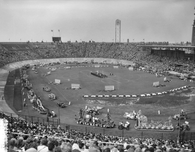 Schwarzes Bild einer großen Menschenmenge, die ein Rennen in einem Stadion beobachtet, mit Fahrzeugen, Zäunen, Pfosten, Flaggen, Türmen, Bäumen und Himmel im Hintergrund.