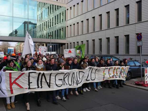 Eine Gruppe von Menschen, die auf einer Straße stehen und ein Transparent mit der Aufschrift 'Kein Grind Lo Feuer' halten, vor dem Hintergrund von Gebäuden, Bäumen, Polen, Schildern, Fahrzeugen und einem klaren blauen Himmel.