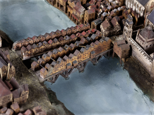 Ein detailliertes Miniaturmodell einer Stadtlandschaft mit einem Fluss, das eine Brücke über das Wasser spannt und zahlreiche Gebäude enthält.