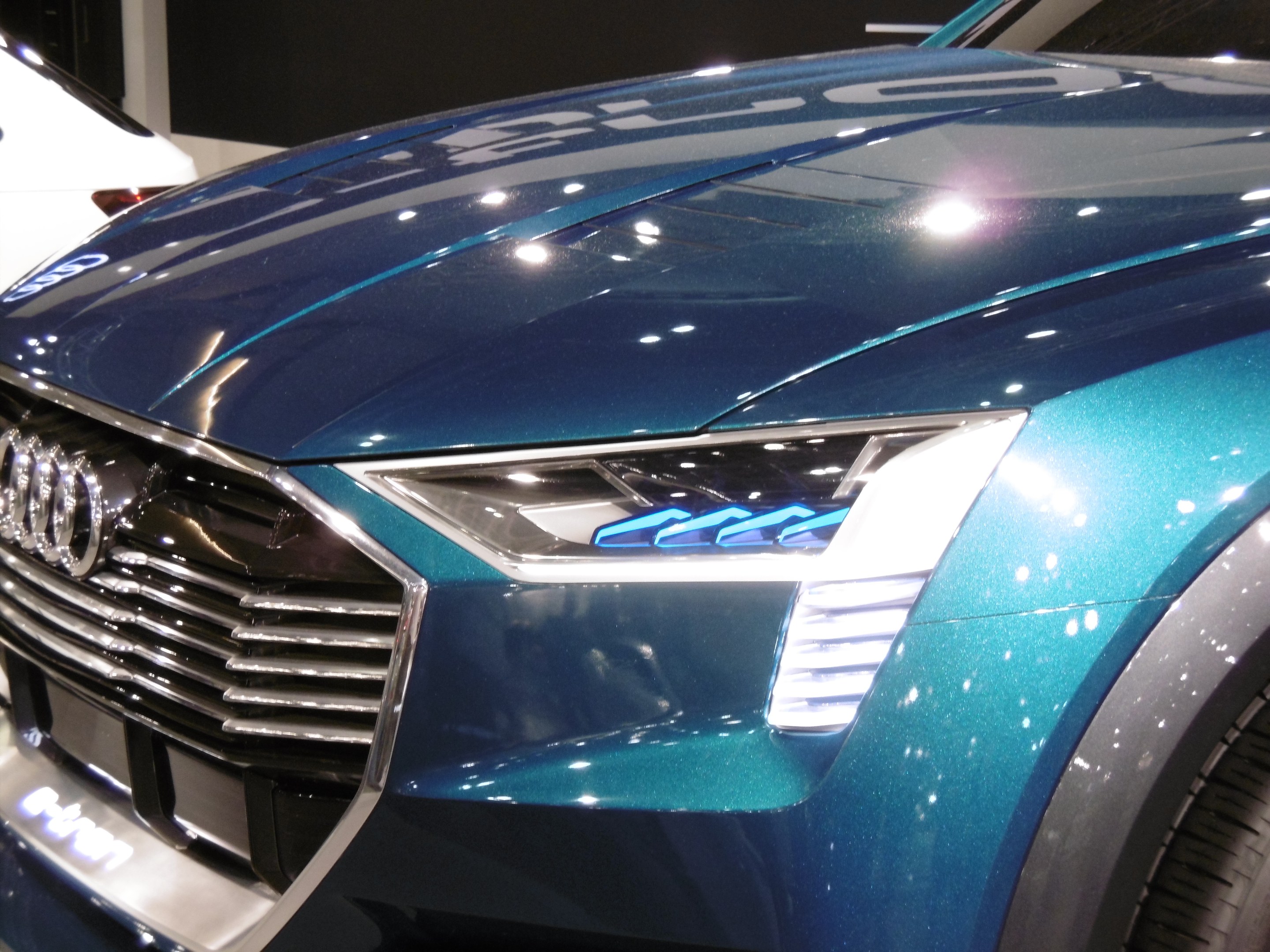 Blauer Audi e-tron Conceptwagen auf einer Automesse ausgestellt, mit einem weißen Auto im Hintergrund und einer schwarzen Wand dahinter.